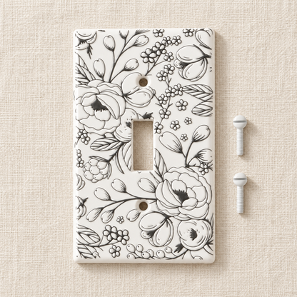 A single-toggle light switch plate cover with a black floral line-art pattern on a white background, shown with two white mounting screws on a textured beige surface.