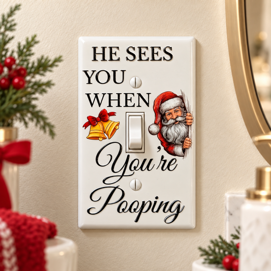 A novelty light switch plate mounted on a beige wall, featuring the text 'HE SEES YOU WHEN You're Pooping' in black lettering, a vintage-style Santa Claus illustration, and a gold bell graphic. 