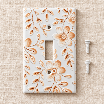 A single-toggle light switch plate cover featuring a white background with a raised floral pattern in warm peach and tan tones, shown on a beige textured surface with two white mounting screws beside it.